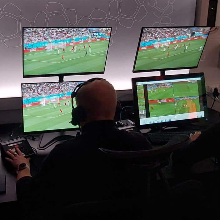 Two people wearing headsets sit at a desk with multiple computer monitors showing a soccer game. A large wall sign reads 'VIDEOKOHTUNIK' in a dark room with technical equipment and a security camera overhead.