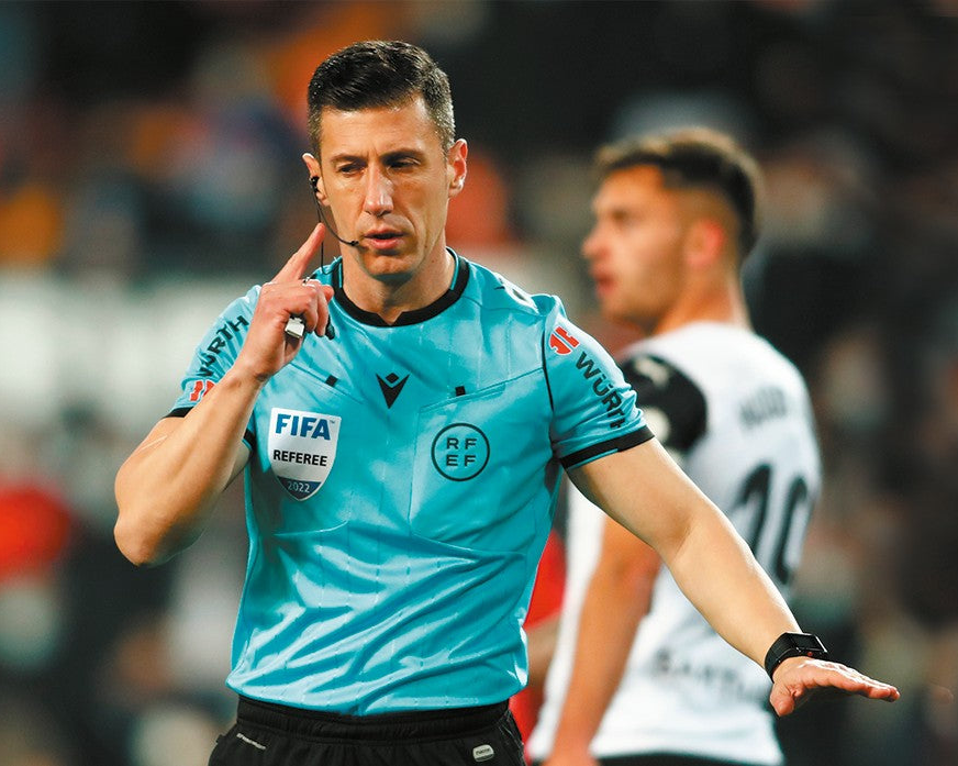 A soccer referee in a light blue FIFA uniform with black shorts, gesturing with his hands on the field, with a blurred stadium crowd in the background.