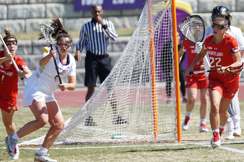 Lacrosse game in progress with players on a field, goal in background