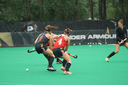 Two female field hockey players in action on a green field with 'Under Armour' branding in the background.